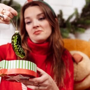 A disappointed woman opening a holiday gift with a pickle ornament inside.